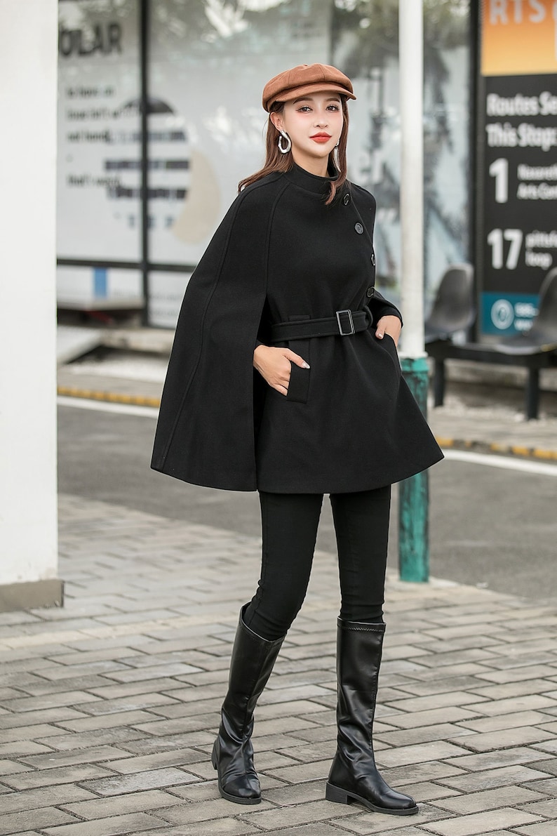May include: A black wool cape with a high collar, a belt, and a button closure. The cape is worn over a black mock neck shirt and black pants. The person is wearing brown boots and a brown newsboy cap.