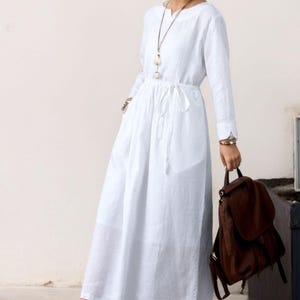 May include: A white linen dress with long sleeves and a drawstring waist. The dress has a v-neckline and a relaxed fit. The model is wearing the dress with brown leather shoes.