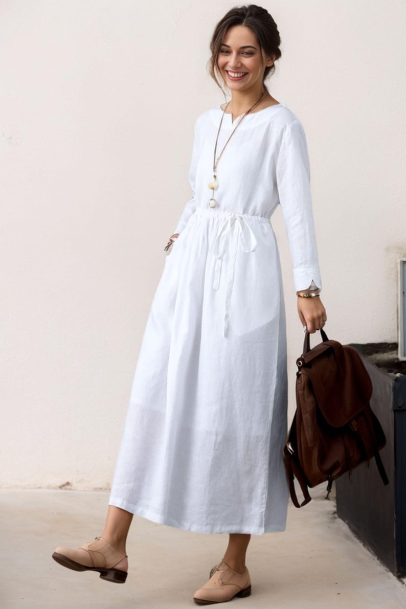 May include: A white linen dress with long sleeves and a drawstring waist. The dress has a v-neckline and a relaxed fit. The model is wearing the dress with brown leather shoes.