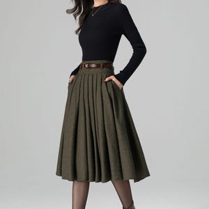 May include: A woman wearing a black long-sleeved top and an olive green pleated skirt with a brown belt. She is also wearing brown boots and black tights. The outfit is a classic, elegant style.