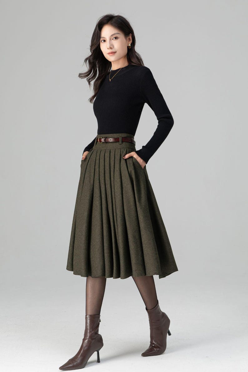 May include: A woman wearing a black long-sleeved top and an olive green pleated skirt with a brown belt. She is also wearing brown boots and black tights. The outfit is a classic, elegant style.