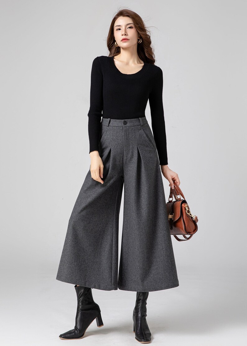 Pu&ograve; includere: Una donna indossa un maglione nero a maniche lunghe e pantaloni larghi grigi. Tiene una borsa di pelle marrone.