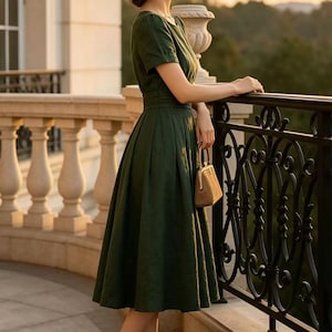 May include: A dark green midi dress with short sleeves and a fitted waist, paired with a tan straw hat, a small tan handbag with a metal frame, and tan high-heeled pumps featuring black pointed toes, worn in a setting with a stone balcony railing.