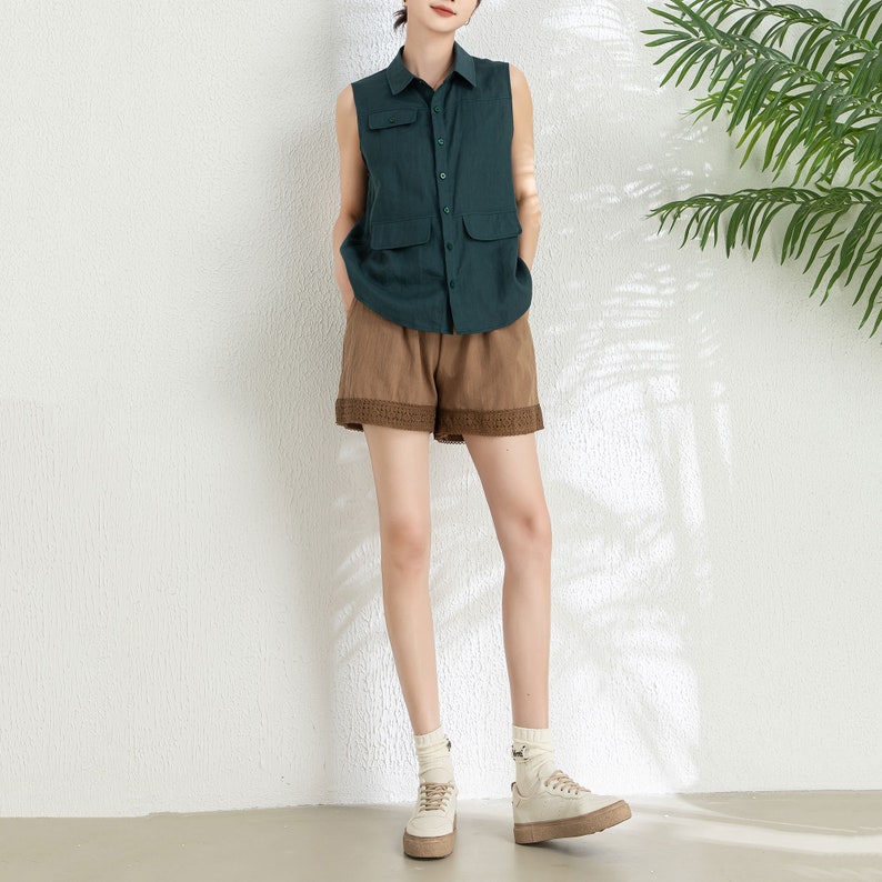 May include: A sleeveless green linen shirt with a button-down collar and two pockets. The shirt is paired with brown shorts that have a lace trim. The model is wearing white sneakers.