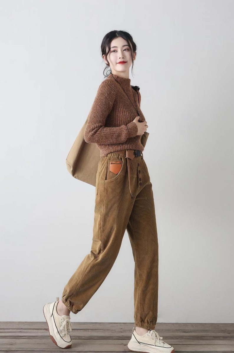 K&ouml;nnte beinhalten: Eine Frau tr&auml;gt einen braunen Rollkragenpullover und eine braune Cordhose mit einem braunen Lederg&uuml;rtel und einer braunen Leder-Patch-Tasche. Sie tr&auml;gt wei&szlig;e Turnschuhe und tr&auml;gt eine braune Papiert&uuml;te.