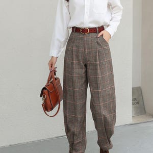K&ouml;nnte beinhalten: Ein wei&szlig;es Hemd mit Knopfleiste und Tasche wird mit einer braunen karierten Hose und einem braunen Lederg&uuml;rtel kombiniert. Eine braune Lederhandtasche und braune Stiefel vervollst&auml;ndigen das Outfit. Die Hose hat ein plissiertes Design. Der Gesamtstil ist klassisch und schick.