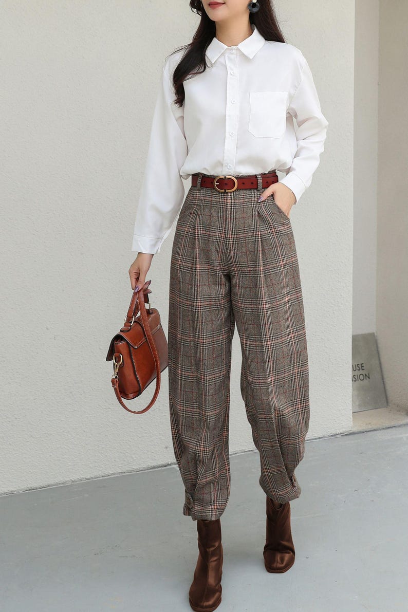 K&ouml;nnte beinhalten: Ein wei&szlig;es Hemd mit Knopfleiste und Tasche wird mit einer braunen karierten Hose und einem braunen Lederg&uuml;rtel kombiniert. Eine braune Lederhandtasche und braune Stiefel vervollst&auml;ndigen das Outfit. Die Hose hat ein plissiertes Design. Der Gesamtstil ist klassisch und schick.