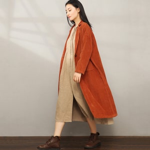 May include: A long, rust-colored corduroy coat with a button closure and pockets. The coat is worn over a beige dress.