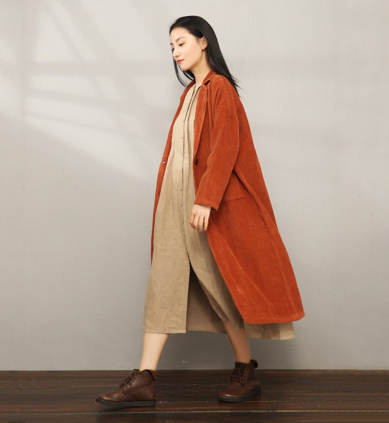 May include: A long, rust-colored corduroy coat with a button closure and pockets. The coat is worn over a beige dress.