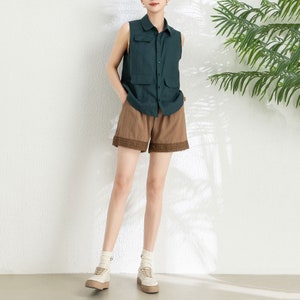 May include: A woman is wearing a dark green sleeveless shirt with a button-up front and two pockets. She is also wearing brown shorts with a lace trim and white sneakers.