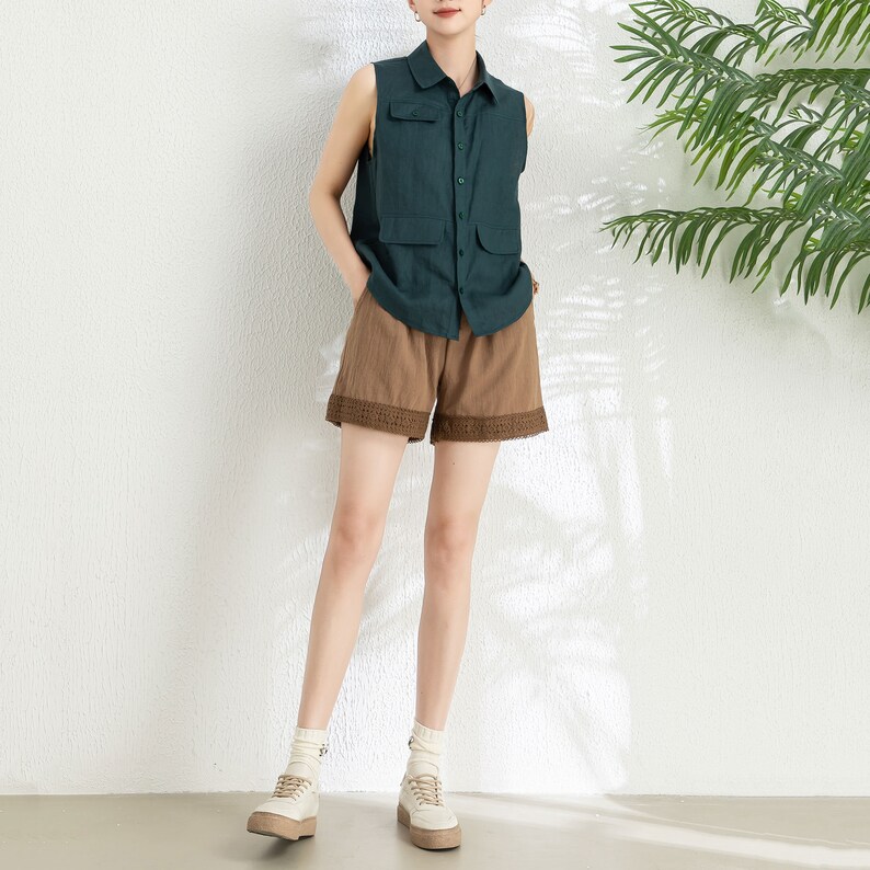 May include: A woman is wearing a dark green sleeveless shirt with a button-up front and two pockets. She is also wearing brown shorts with a lace trim and white sneakers.