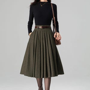 May include: A woman's outfit featuring a black long-sleeved top and a pleated olive green midi skirt. The outfit is accessorized with a brown belt, a brown shoulder bag, and brown ankle boots. The overall style is classic and elegant.