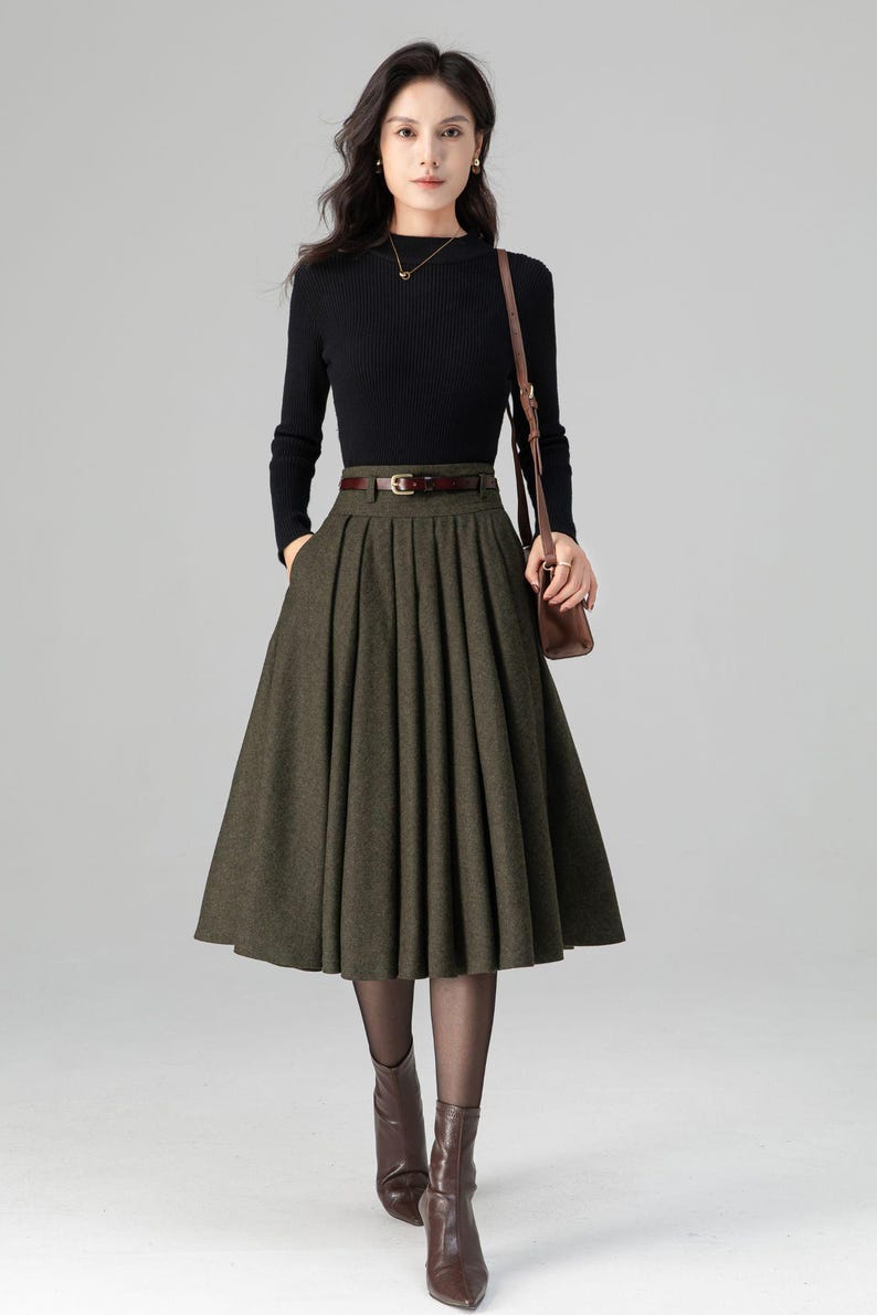 May include: A woman's outfit featuring a black long-sleeved top and a pleated olive green midi skirt. The outfit is accessorized with a brown belt, a brown shoulder bag, and brown ankle boots. The overall style is classic and elegant.