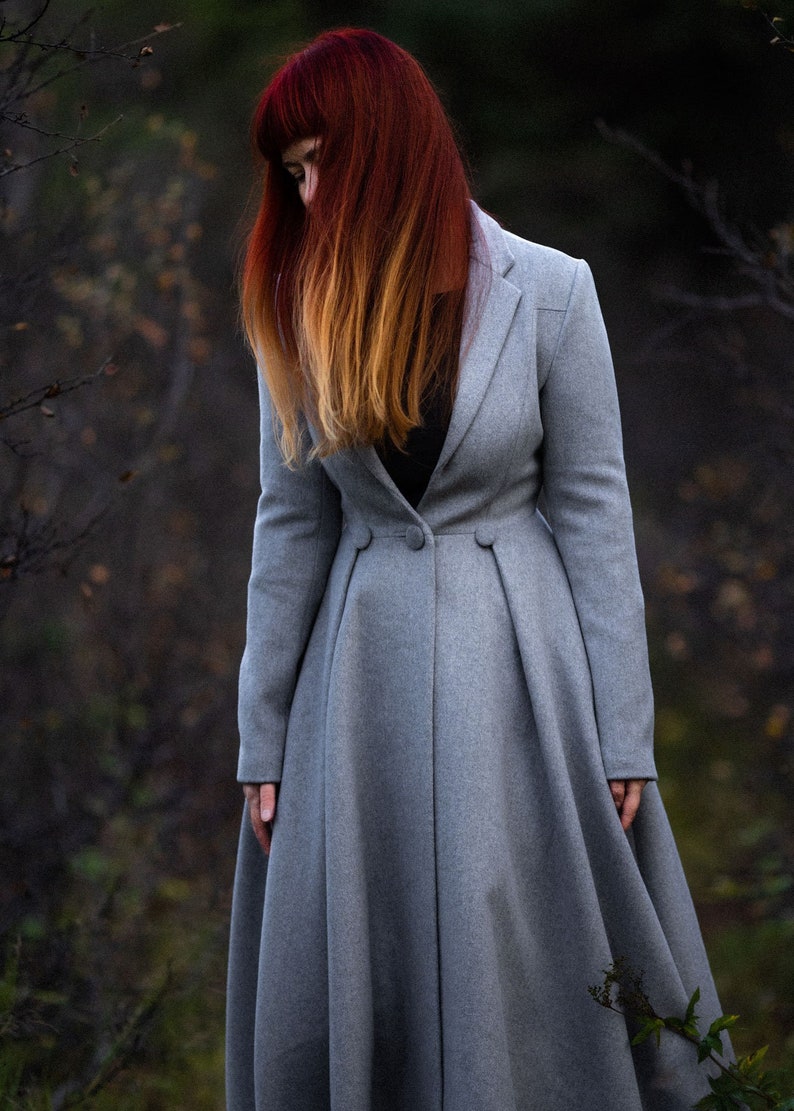 May include: A long, light gray coat with a tailored fit. The coat features a button closure and a flared skirt. The person in the image has red hair with blonde highlights. The coat is a classic style, suitable for various occasions.