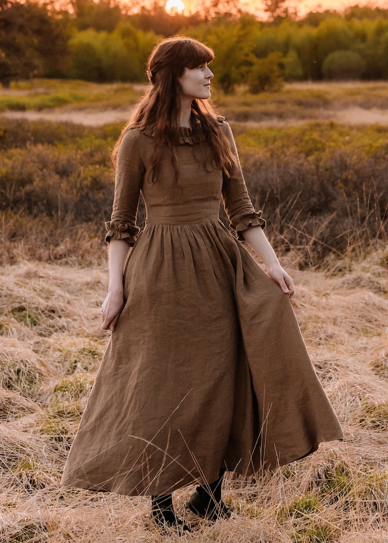 Puede incluir: Una mujer con un vestido largo de lino marr&oacute;n se encuentra en un campo de hierba alta. El vestido tiene un cuello y mangas con volantes, y la mujer mira hacia un lado. El sol se pone en el fondo, proyectando un c&aacute;lido resplandor en la escena.