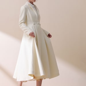 May include: A white long-sleeved coat with a flared skirt and a single button closure. The coat is worn by a person wearing a straw hat with a brown band and white heels.