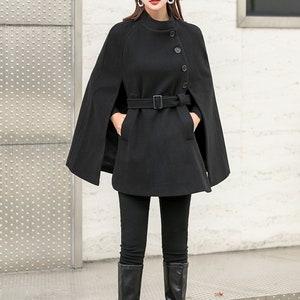 May include: A black wool cape with a belt and buttons. The cape has a wide collar and is worn over a black turtleneck shirt and black pants. The person is wearing black knee-high boots and a brown newsboy cap.