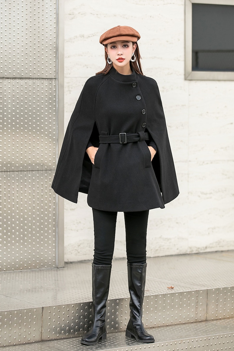 May include: A black wool cape with a belt and buttons. The cape has a wide collar and is worn over a black turtleneck shirt and black pants. The person is wearing black knee-high boots and a brown newsboy cap.