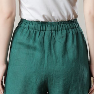 May include: Close-up of emerald green linen shorts with an elastic waistband. The shorts are paired with a white top. The fabric appears to be lightweight and breathable, suggesting a casual style.
