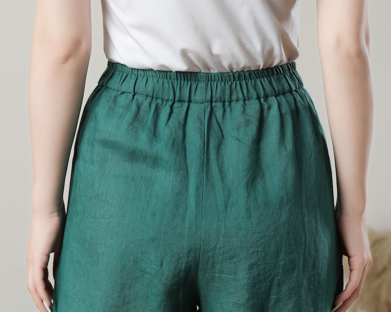 May include: Close-up of emerald green linen shorts with an elastic waistband. The shorts are paired with a white top. The fabric appears to be lightweight and breathable, suggesting a casual style.
