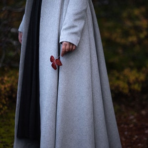 May include: A light gray, long wool coat with a flared skirt and two large buttons. A person holds a small, red leaf. The coat is paired with a black dress. The background is blurred with fall colors.