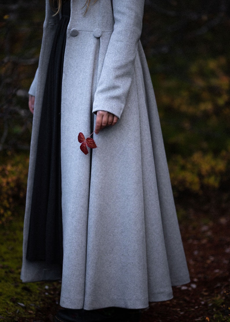 May include: A light gray, long wool coat with a flared skirt and two large buttons. A person holds a small, red leaf. The coat is paired with a black dress. The background is blurred with fall colors.
