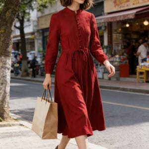 Könnte beinhalten: Ein knielanges, rotes Kleid mit langen Ärmeln, Knopfleiste und Bindegürtel. Dazu werden braune Schuhe getragen. Eine Person trägt eine braune Papiereinkaufstasche, während sie auf einem Bürgersteig geht.