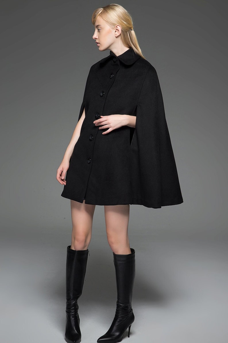May include: A black wool cape with a collar and button fastening. The cape is worn over a black dress and the model is wearing black knee-high boots.
