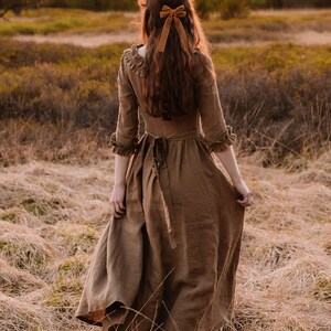 Puede incluir: Una mujer con un vestido de lino marr&oacute;n y un lazo de terciopelo marr&oacute;n en el pelo camina por un campo de hierba alta.