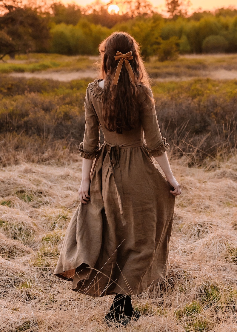 Puede incluir: Una mujer con un vestido de lino marr&oacute;n y un lazo de terciopelo marr&oacute;n en el pelo camina por un campo de hierba alta.