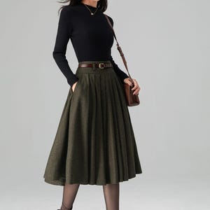 May include: A woman's outfit featuring a black ribbed long-sleeve top, a dark green pleated skirt, and brown leather accessories. The skirt is knee-length and has a subtle pattern. The outfit is completed with brown ankle boots and a matching shoulder bag.