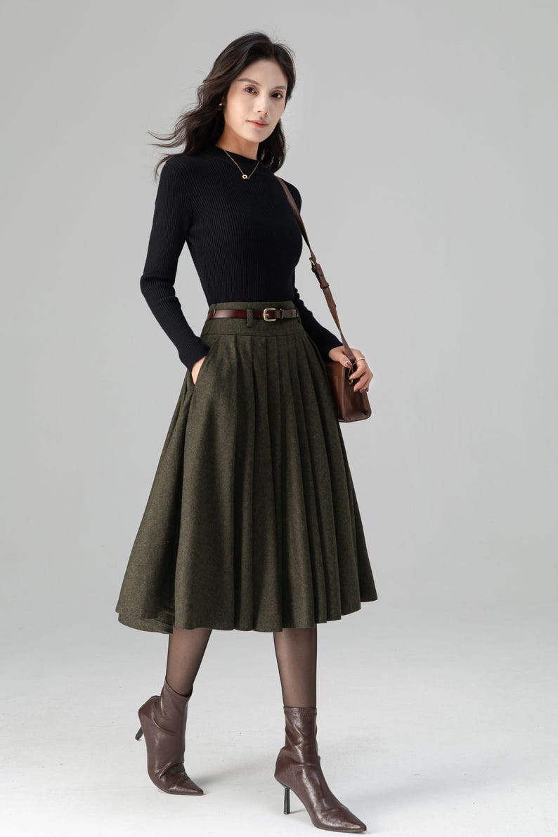 May include: A woman's outfit featuring a black ribbed long-sleeve top, a dark green pleated skirt, and brown leather accessories. The skirt is knee-length and has a subtle pattern. The outfit is completed with brown ankle boots and a matching shoulder bag.