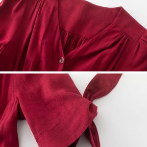 May include: A close-up of a burgundy linen dress with a V-neckline and button closure. The dress has short sleeves with a tie detail at the cuff.