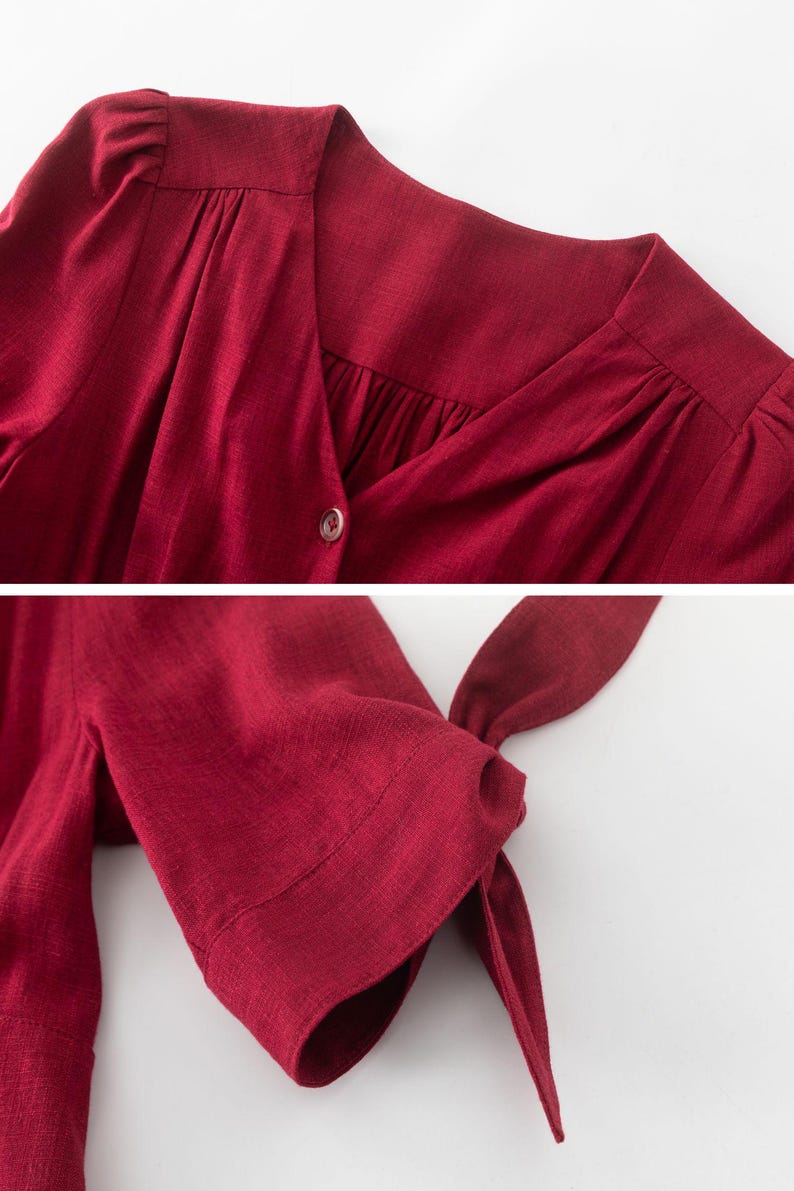 May include: A close-up of a burgundy linen dress with a V-neckline and button closure. The dress has short sleeves with a tie detail at the cuff.