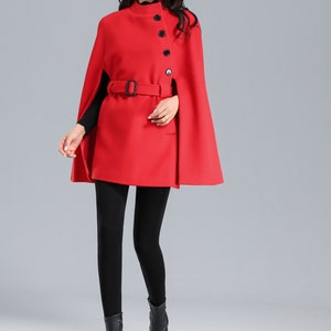 May include: A red wool cape with a high collar, a belt, and four black buttons. The cape has a slit on each side. The model is wearing black leggings and black boots.