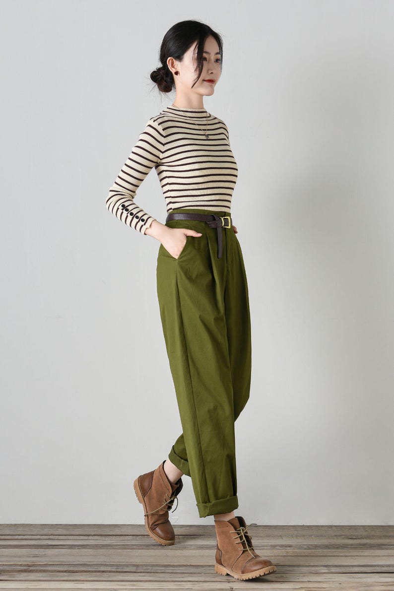 Op de afbeelding: Een vrouw draagt een gestreept, cr&egrave;me en bruin, lang mouwen shirt met een bruine leren riem en een olijfgroene broek. Ze draagt ​​bruine leren enkellaarsjes.