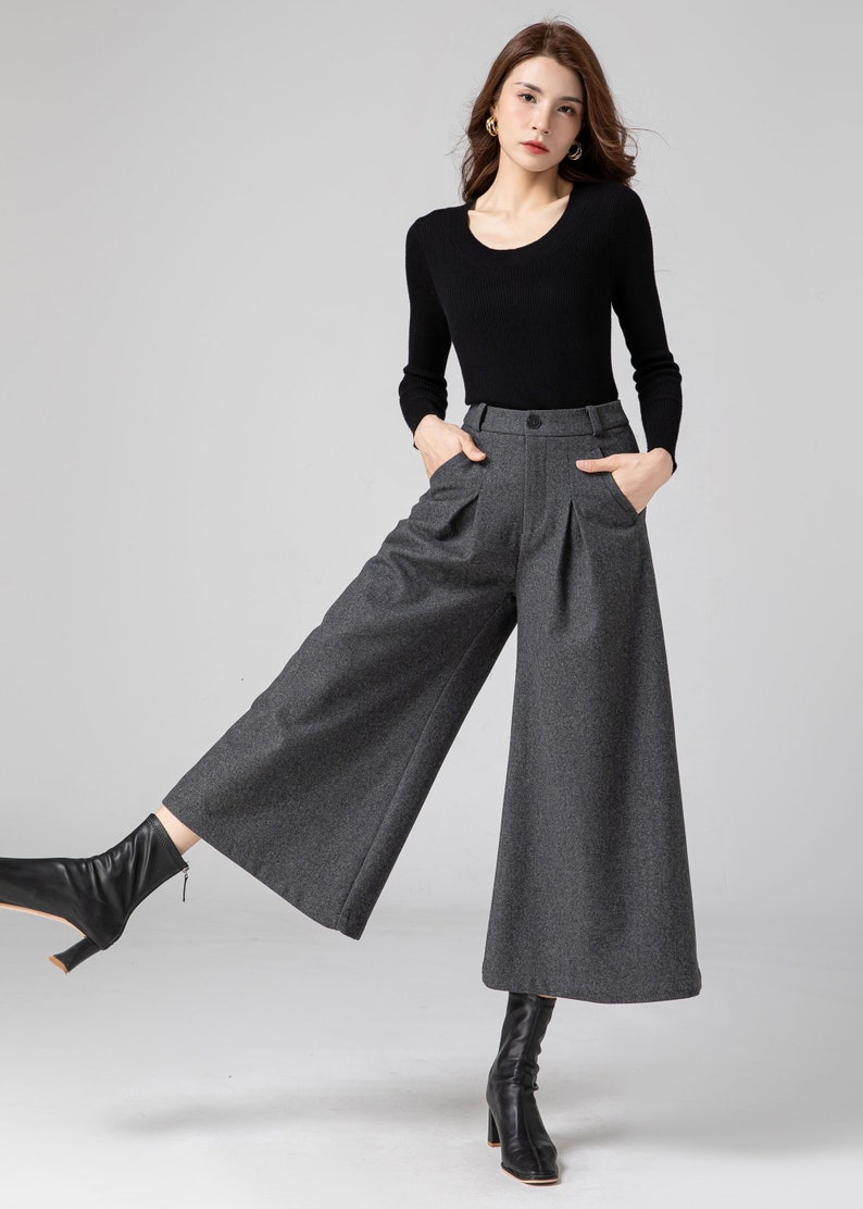 Pu&ograve; includere: Una donna indossa un maglione nero a maniche lunghe e pantaloni larghi grigi con pieghe. Indossa stivaletti neri alla caviglia.