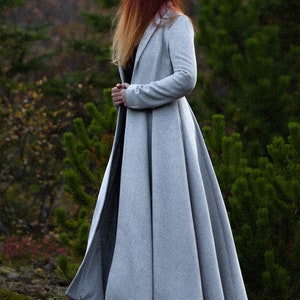 May include: A long, light gray coat with a flared skirt, worn outdoors. The coat has a classic design and is made of a soft material. The person in the coat has long red hair. The coat is suitable for cooler weather.
