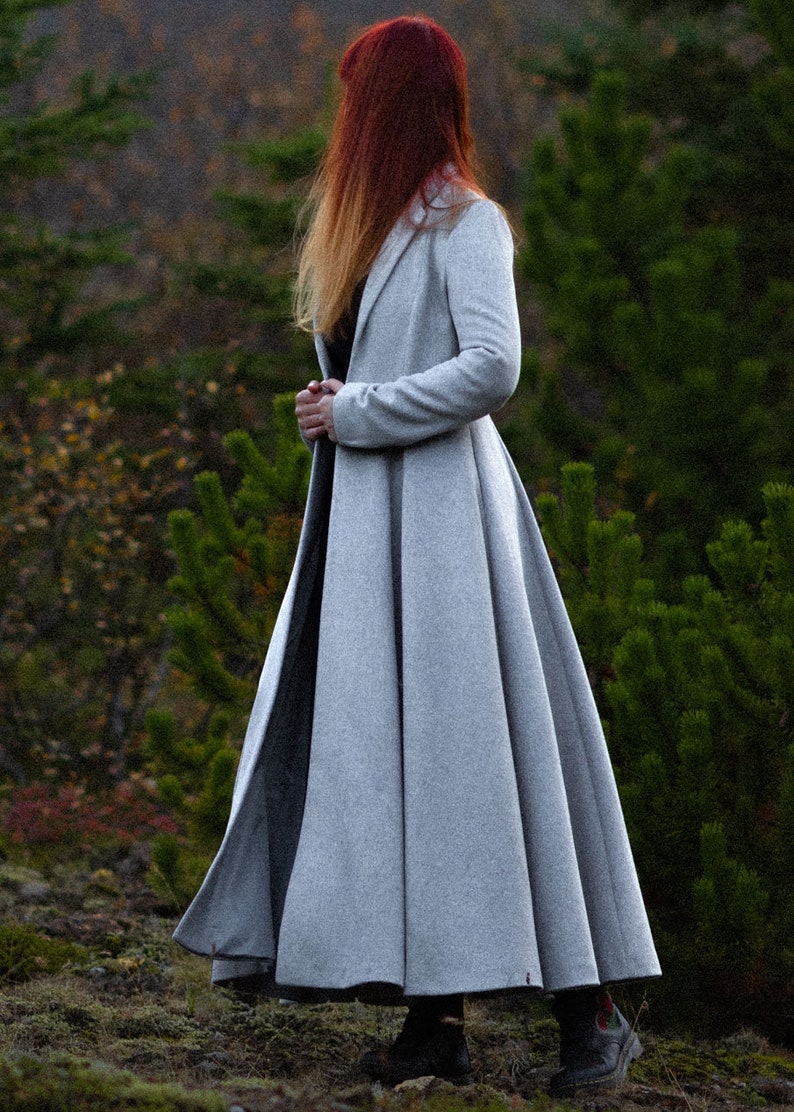 May include: A long, light gray coat with a flared skirt, worn outdoors. The coat has a classic design and is made of a soft material. The person in the coat has long red hair. The coat is suitable for cooler weather.