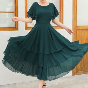 May include: A dark green midi dress with a tiered skirt and flutter sleeves. The dress has a fitted waist and a flowing, layered skirt. The model is wearing black sandals. The dress is made of a lightweight fabric.