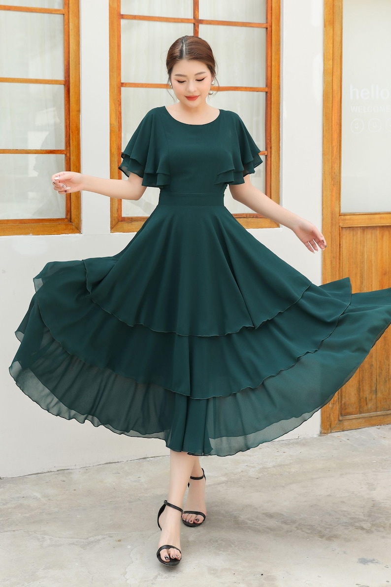 May include: A dark green midi dress with a tiered skirt and flutter sleeves. The dress has a fitted waist and a flowing, layered skirt. The model is wearing black sandals. The dress is made of a lightweight fabric.