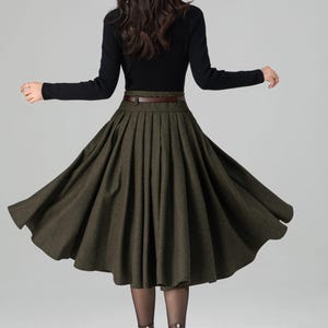 May include: A woman's outfit featuring a black long-sleeve top and a dark green pleated skirt. The skirt is cinched at the waist with a brown belt. Brown heeled boots complete the look. The outfit is suitable for fall or winter.