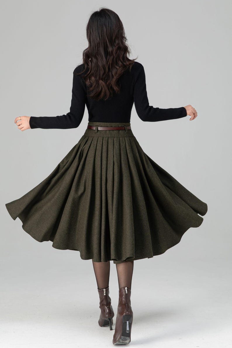 May include: A woman's outfit featuring a black long-sleeve top and a dark green pleated skirt. The skirt is cinched at the waist with a brown belt. Brown heeled boots complete the look. The outfit is suitable for fall or winter.