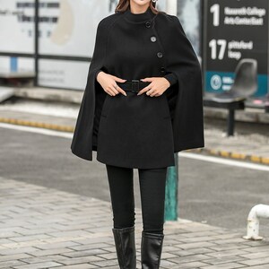 May include: A black cape with a high neck and a single button closure. The cape has a belt and is worn with black leggings and black knee-high boots. The person is wearing a brown newsboy cap.