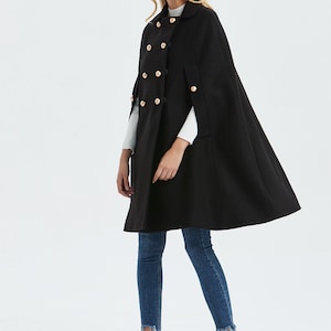 May include: A black double-breasted cape with gold buttons. The cape has a collar and is knee-length. The model is wearing blue jeans and black ankle boots.