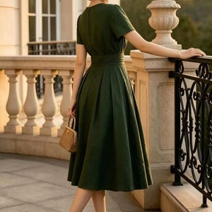 May include: A dark green midi dress with short sleeves, a fitted waist, and a pleated skirt. The outfit includes a tan straw hat with a black band, tan high-heeled pumps, and a small tan handbag with a gold-tone metal frame and clasp closure.