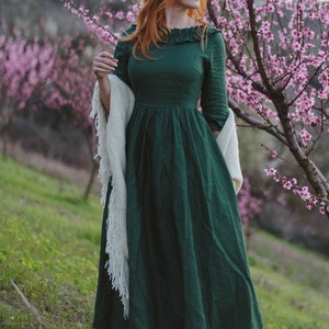 Puede incluir: Una mujer con el pelo largo y rojo lleva un vestido de lino verde con una chalina blanca y botas negras de pie en un campo de &aacute;rboles con flores rosas.