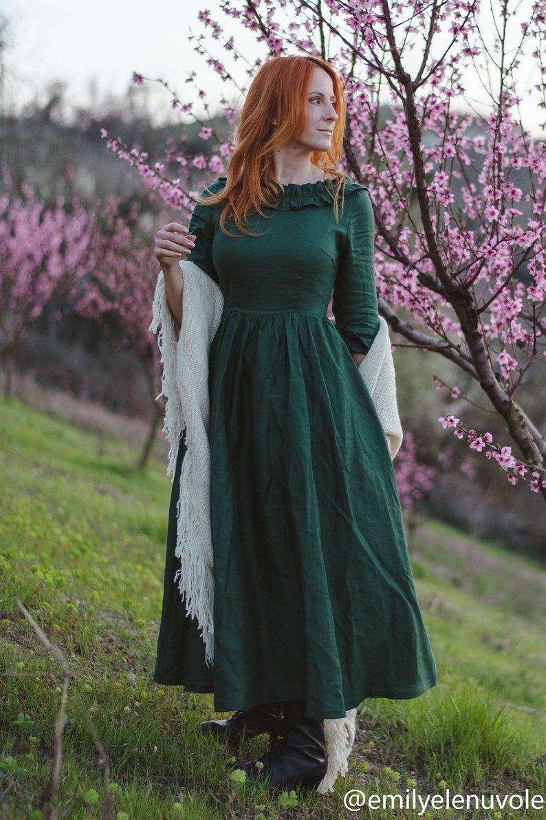 Puede incluir: Una mujer con el pelo largo y rojo lleva un vestido de lino verde con una chalina blanca y botas negras de pie en un campo de &aacute;rboles con flores rosas.
