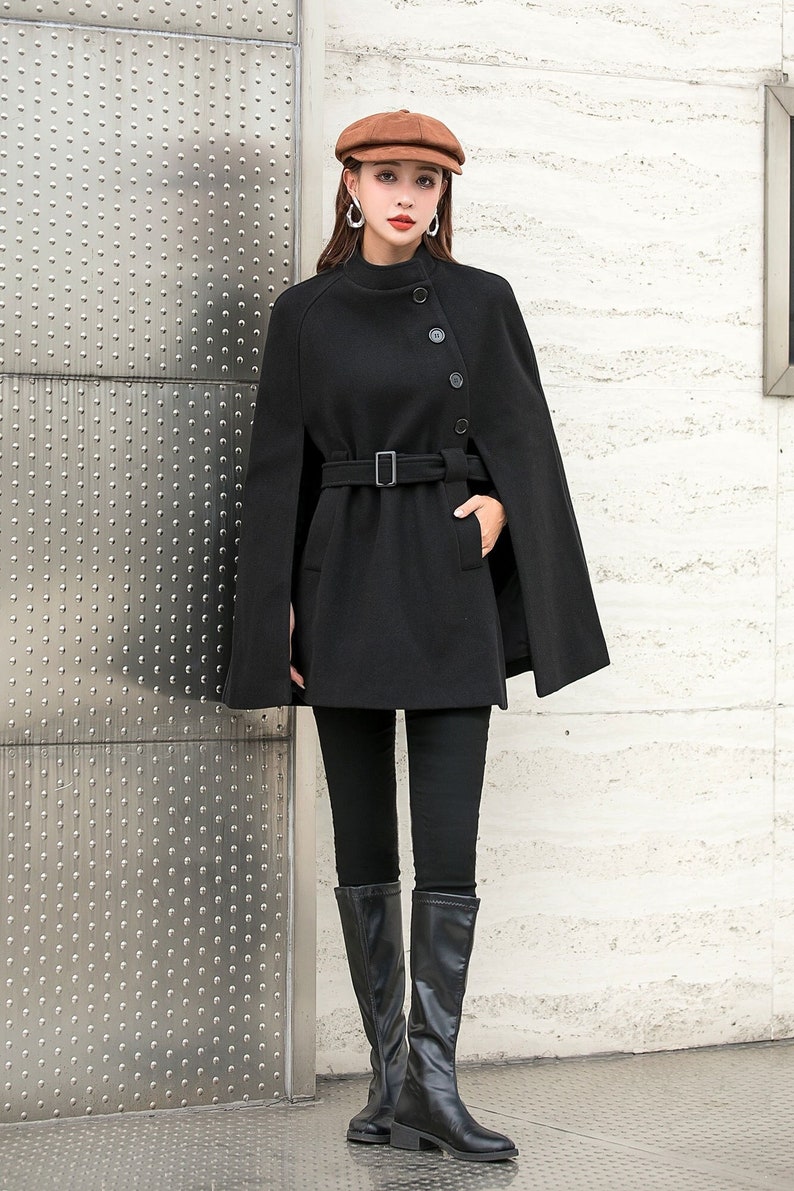 May include: A black cape coat with a belt and buttons. The coat has a high neck and long sleeves. The model is wearing black pants and black boots.