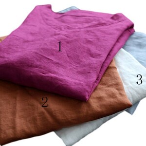 May include: Four folded linen shirts in different colors: a deep magenta, a warm brown, a soft white, and a light blue. Each shirt has a simple, round neckline. The shirts are stacked on top of each other, showcasing the variety of colors.